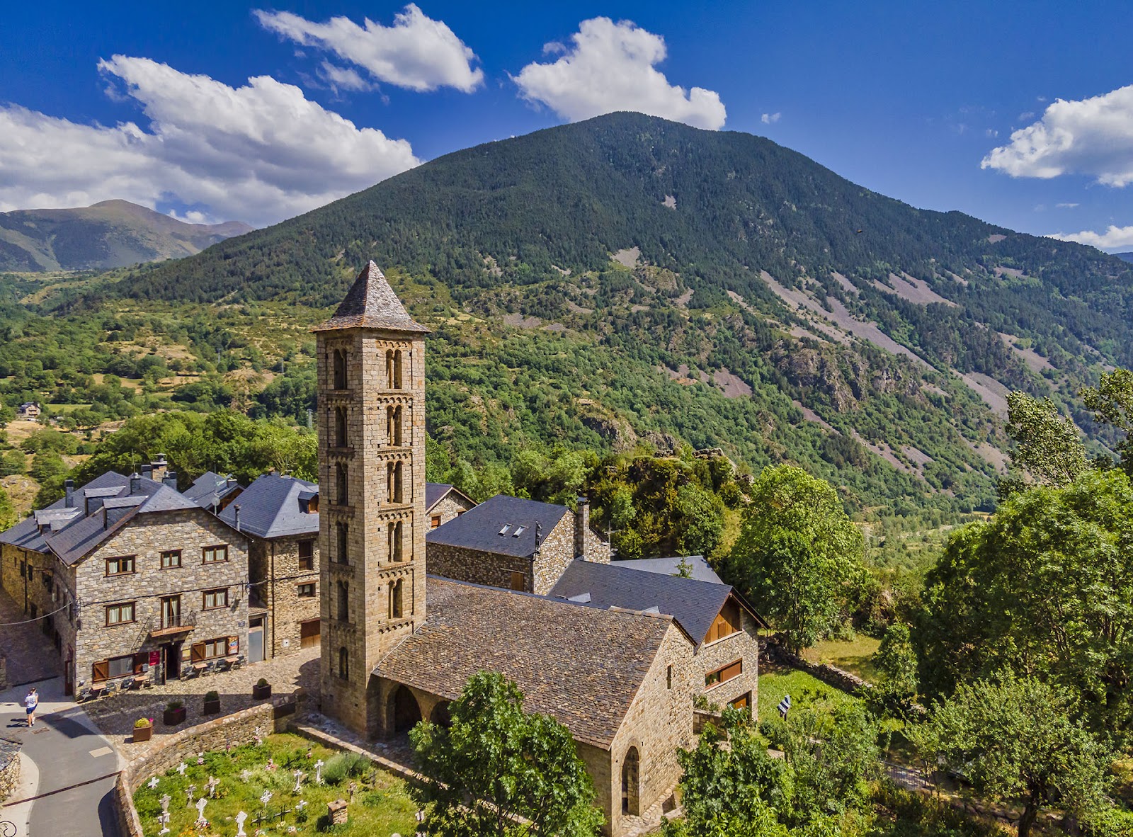 Vista aèria de l’església de Santa Eulàlia d’Erill la Vall amb algunes cases del poble al darrere i les muntanyes de fons.