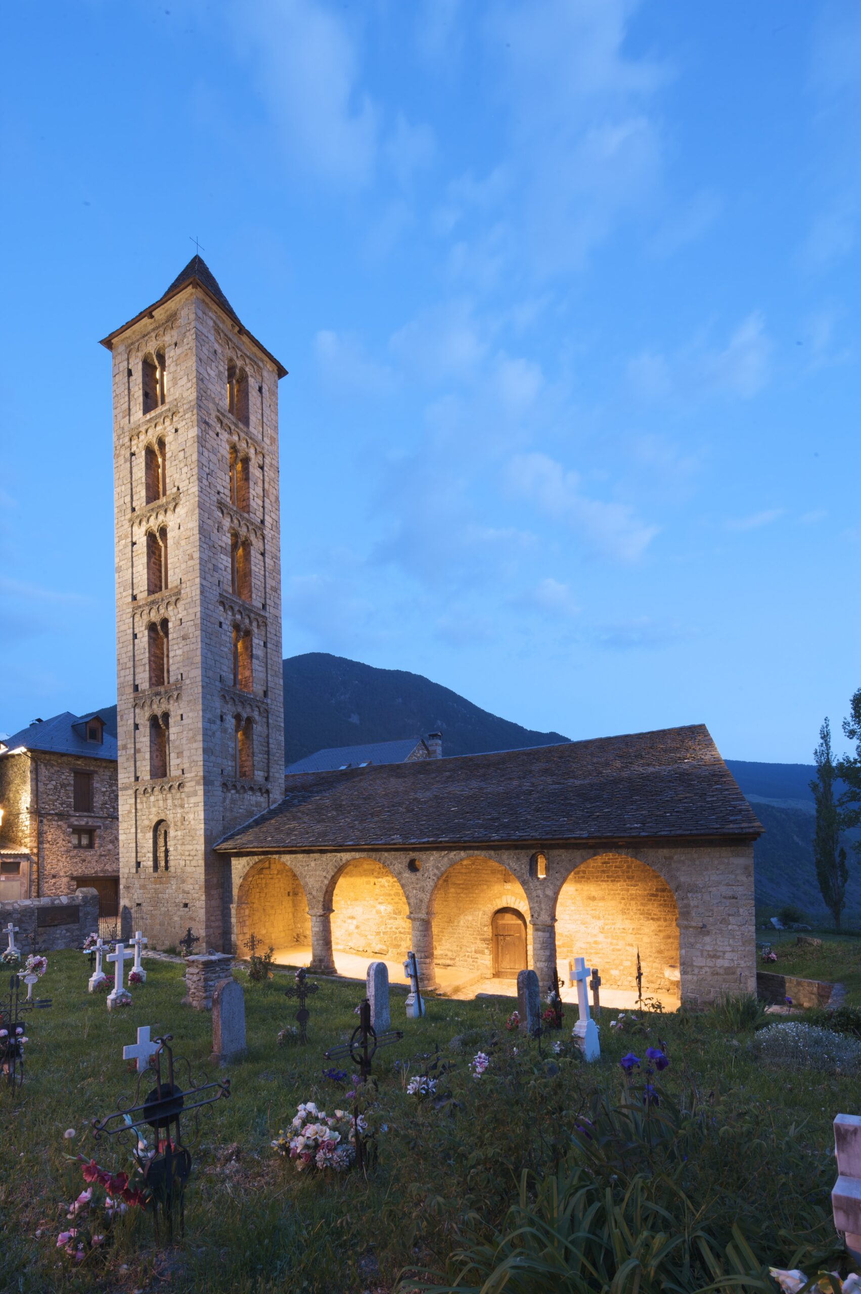 Exterior de l’església de Santa Eulàlia d’Erill la Vall al capvespre, amb el porxo il·luminat i el cementiri al davant.