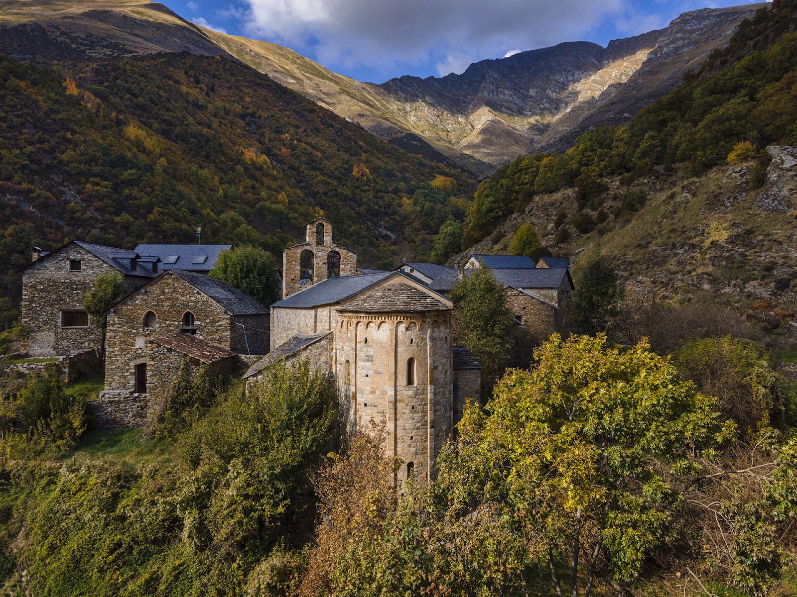 Vista posterior de l’església de Santa Maria de Cardet envoltada de vegetació, amb l’absis construït seguint el desnivell de la muntanya.