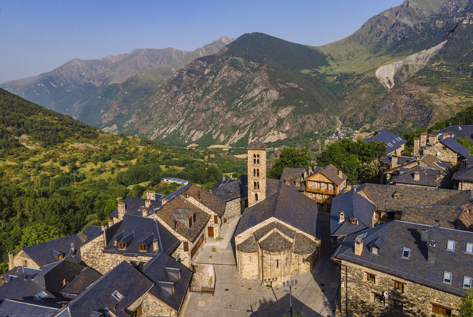 Vista aèria de l’església de Santa Maria de Taüll envoltada per diverses cases del poble i les muntanyes al fons.