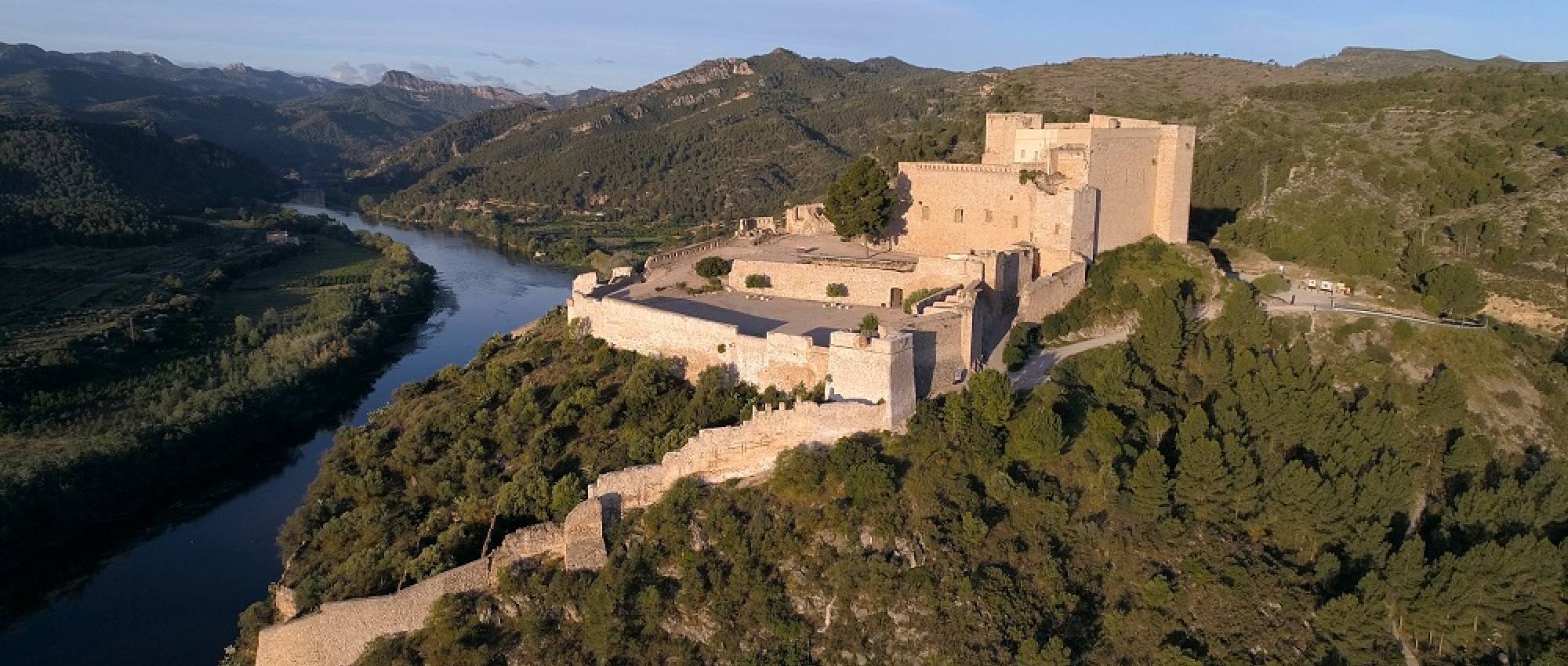 Vista aèria del castell de Miravet amb petits turons al darrera i el riu Ebre a l’esquerra.