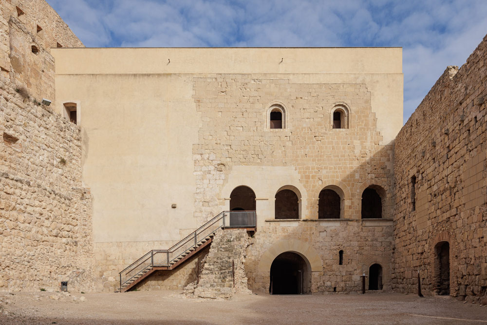Vista general del pati d’armes del castell amb la façana de l’edifici noble al fons. A la planta baixa de l’edifici hi ha les portes d’entrada al celler i magatzem; a la primera planta, les quatre finestres de la galeria; i a la segona planta, les dues finestres de la sala del comanador.