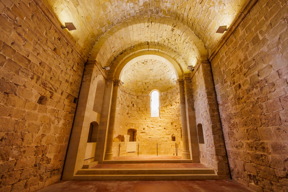 Vista de l’altar de l’església amb la porta d’accés a la torre del tresor a l’esquerra. S’accedeix a l’altar mitjançant uns graons. Al mur de l’absis hi ha una finestra allargada que deixa entrar la llum del matí.